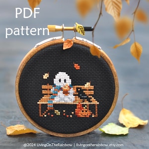 May include: A cross-stitch pattern featuring a ghost wearing a scarf and holding a pumpkin, sitting on a bench with a black cat and a pumpkin. The pattern is in a wooden embroidery hoop, with autumn leaves in the background. The text "PDF pattern" is at the top of the image.