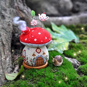 以下が含まれることがあります： 赤い白のきのこ型の針山で、小さな家のデザインと、その上にティーポットとティーカップが乗っています。針山は木製の台座の上に置かれ、緑の苔に囲まれています。