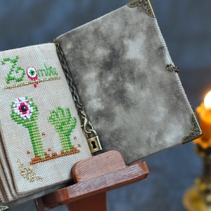 May include: A cross-stitched book with a zombie theme. The cover is a mottled gray, and the pages display green zombie hands and the word "Zombie" in green. The book is held open by a wooden stand, with a lit candle in the background.