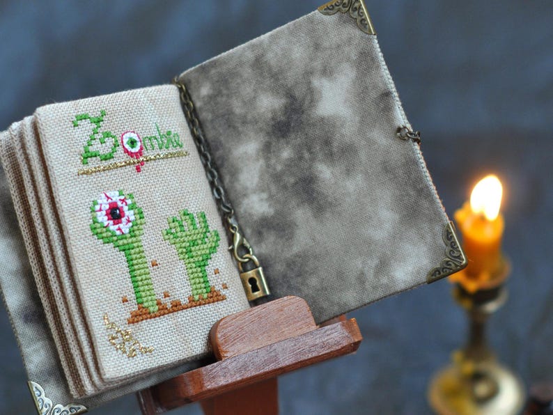 May include: A cross-stitched book with a zombie theme. The cover is a mottled gray, and the pages display green zombie hands and the word "Zombie" in green. The book is held open by a wooden stand, with a lit candle in the background.