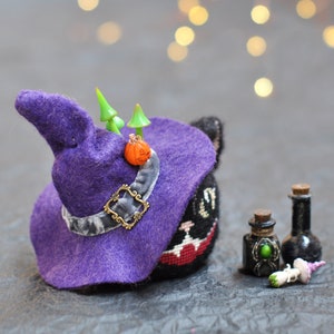 May include: A black cat wearing a purple witch's hat, a small orange pumpkin and green mushrooms. The cat is wearing a black and white striped shirt and has a red and white striped collar. The cat is sitting on a grey surface.