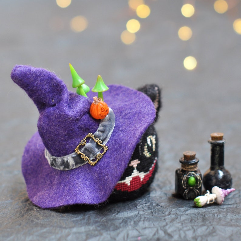 May include: A black cat wearing a purple witch's hat, a small orange pumpkin and green mushrooms. The cat is wearing a black and white striped shirt and has a red and white striped collar. The cat is sitting on a grey surface.