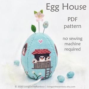May include: A light blue egg-shaped pincushion with a cross-stitch design featuring an egg house with penguins. The pincushion has a red roof and a ladder. The text "Egg House PDF pattern no sewing machine required" is visible.