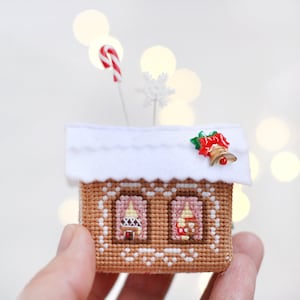 Op de afbeelding: Een gingerbreadhuis ornament met een wit dak, een rode snoepstok en een rode bel met groene hulstbladeren. Het huis heeft twee ramen met roze gordijnen en bruine trim.