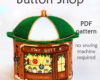 Könnte beinhalten: Ein Kreuzstichmuster für einen Knopfladenschmuck. Der Schmuck ist ein kleines, rundes Gebäude mit einem grünen Dach und einem Schild mit der Aufschrift "Tiny Button". Das Muster ist als PDF-Download erhältlich und erfordert keine Nähmaschine.