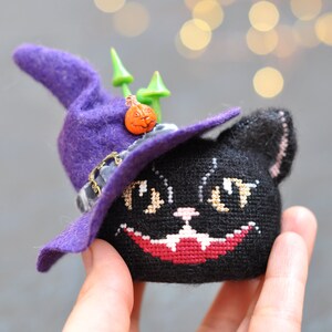 May include: A black cat wearing a purple witch's hat with a small orange pumpkin decoration. The cat has a stitched-on face with white eyes and a red mouth.