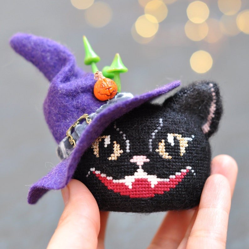 May include: A black cat wearing a purple witch's hat with a small orange pumpkin decoration. The cat has a stitched-on face with white eyes and a red mouth.