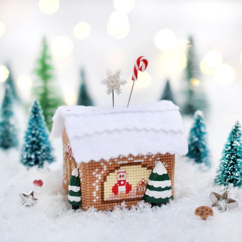 Op de afbeelding: Een wit peperkoekhuis met een rode snoepstok op de top, omringd door miniatuur dennenbomen en een sneeuwvlok. Het peperkoekhuis heeft een wit dak en een peperkoekontwerp aan de voorkant.