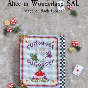 May include: A cross-stitch project titled Alice in Wonderland SAL stage 3: Back Cover. The rectangular piece features a light blue background with the text "curiouser and curiouser" in purple, a red mushroom, and green foliage. It has gold metal corners.