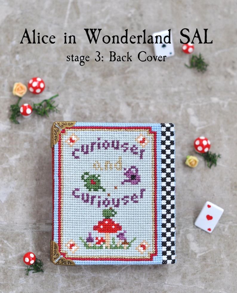May include: A cross-stitch project titled Alice in Wonderland SAL stage 3: Back Cover. The rectangular piece features a light blue background with the text "curiouser and curiouser" in purple, a red mushroom, and green foliage. It has gold metal corners.