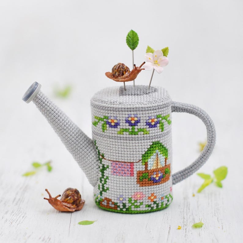 K&ouml;nnte beinhalten: Eine graue Keramik-Gie&szlig;kanne mit einem Kreuzstich-Design eines Hauses mit einem Blumengarten und einer W&auml;scheleine. Zwei kleine braune Schnecken befinden sich auf der Kanne und auf dem Tisch.