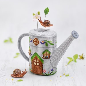 K&ouml;nnte beinhalten: Eine graue Gie&szlig;kanne mit einem Kreuzstich-Design eines Hauses mit einer braunen T&uuml;r und einem gr&uuml;nen Dach. Auf der Kanne befinden sich zwei kleine braune Schnecken und eine wei&szlig;e Blume mit einer braunen Schnecke oben drauf.