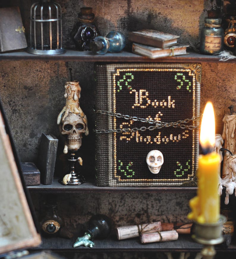 May include: A shelf display featuring a skull candle, a book titled "Book of Shadows", and various potion bottles. The book is secured with a chain and has a skull emblem. Other items include small books and a birdcage.