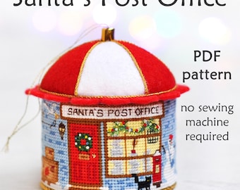 May include: A red and white felt Christmas ornament shaped like a Santa's Post Office building. The ornament is decorated with a wreath, a mailbox, and a black cat. The text "Santa's Post Office" is printed on the front of the ornament. The ornament is a PDF pattern for a no-sew project.