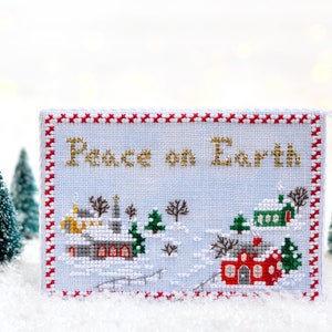 May include: A white cross-stitch ornament with a red and white border. The ornament features a winter scene with houses and the text "Peace on Earth".