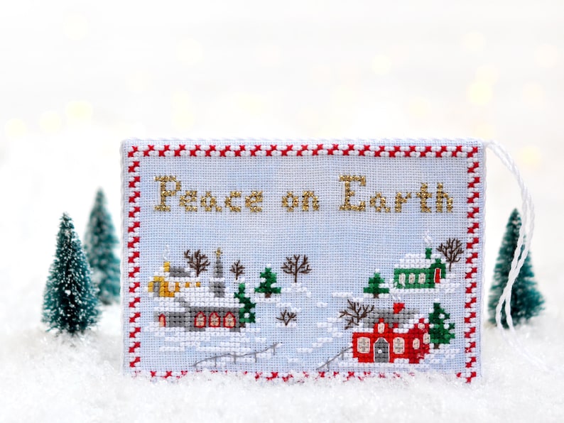May include: A white cross-stitch ornament with a red and white border. The ornament features a winter scene with houses and the text "Peace on Earth".
