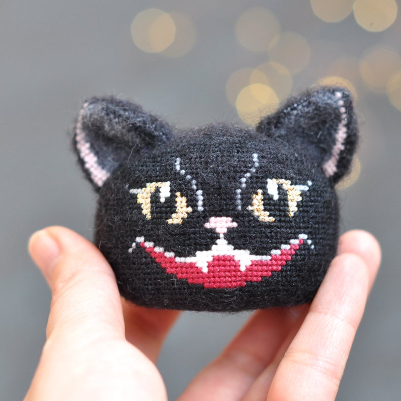 May include: Black plush cat with embroidered yellow eyes, a pink nose, and a red stitched mouth. The cat is grinning with sharp teeth.