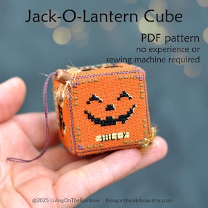May include: An orange, cross-stitched Jack-O-Lantern cube with a smiling face and the word "BOO" stitched on the front. The cube has a purple string attached for hanging. The text "Jack-O-Lantern Cube" and "PDF pattern no experience or sewing machine required" are visible.