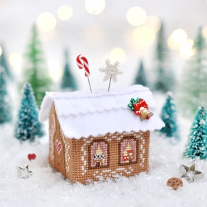 Op de afbeelding: Een naaldenkussen in de vorm van een peperkoekhuis met een wit dak, een rode snoepstok en een sneeuwvlok bovenop. Het naaldenkussen is omringd door miniatuur dennenbomen en een wit besneeuwd oppervlak.