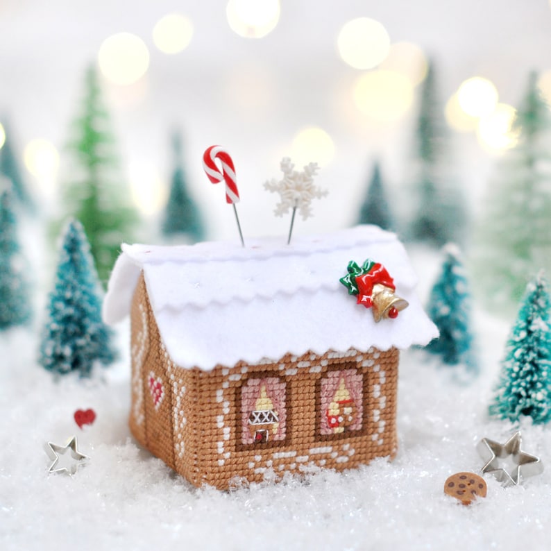 Op de afbeelding: Een naaldenkussen in de vorm van een peperkoekhuis met een wit dak, een rode snoepstok en een sneeuwvlok bovenop. Het naaldenkussen is omringd door miniatuur dennenbomen en een wit besneeuwd oppervlak.