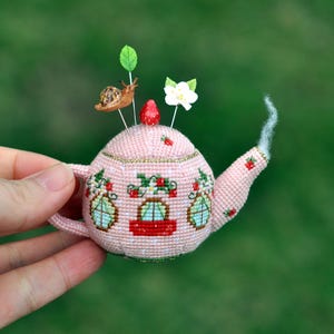 May include: A pink crocheted teapot pincushion with a red strawberry, a white flower, and a snail on top. The teapot has three windows and a red stripe around the middle.