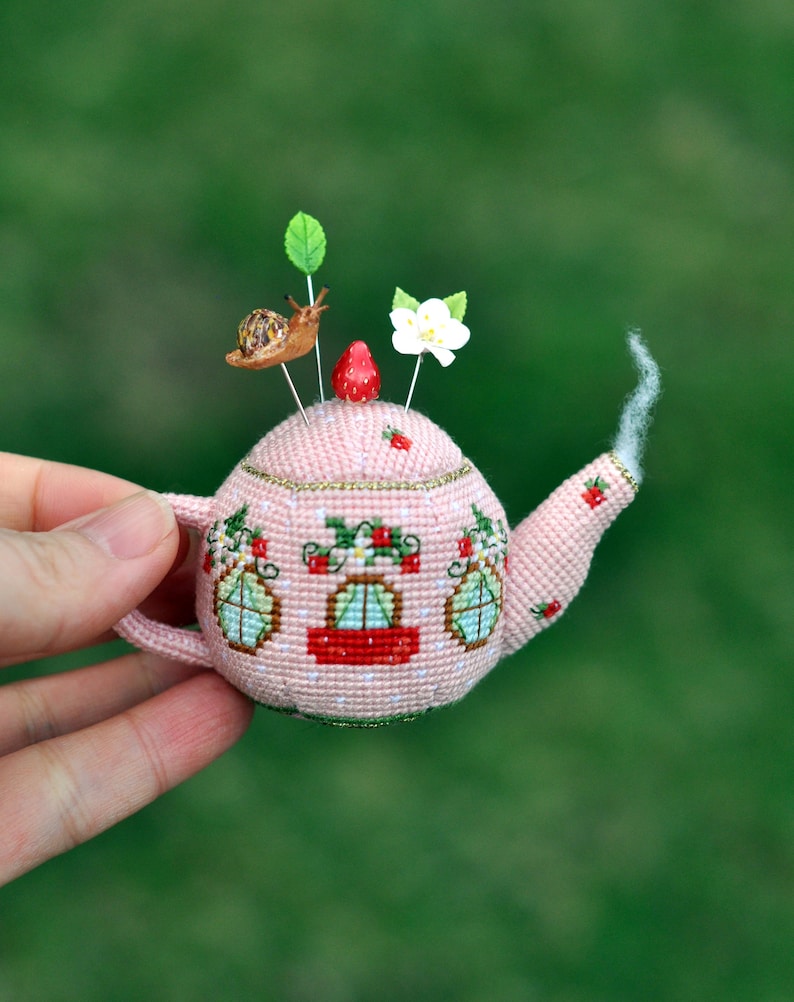 May include: A pink crocheted teapot pincushion with a red strawberry, a white flower, and a snail on top. The teapot has three windows and a red stripe around the middle.