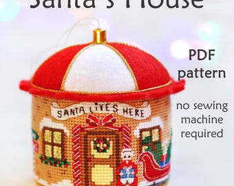 May include: A red and white felt Christmas ornament shaped like a house with a sign that reads "Santa Lives Here". The ornament is decorated with a wreath, a window, and a small Santa Claus figure. The ornament is a PDF pattern for a no-sew project.