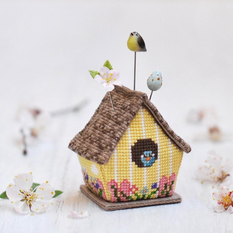May include: A yellow and brown birdhouse pincushion with a brown roof, a blue bird in the window, and a pink flower on top. The pincushion is decorated with pink tulips and green leaves.