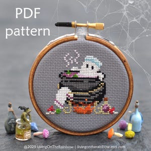 May include: A cross-stitch pattern in a wooden embroidery hoop. The design features a cartoon ghost in a cauldron, with the text "PDF pattern" at the top. The ghost has a blue cap and is surrounded by steam. The cauldron is black and orange.