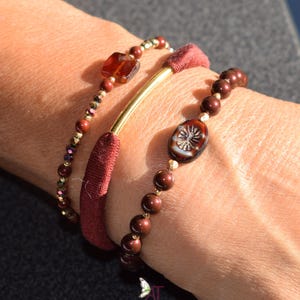 Könnte beinhalten: Drei Armbänder in Braun- und Rottönen. Eines hat kleine, runde Perlen und einen quadratischen, bernsteinfarbenen Stein. Ein weiteres hat einen goldfarbenen Stab und ein bordeauxfarbenes Stoffband. Das dritte Armband hat braune Perlen und eine dekorative ovale Perle.