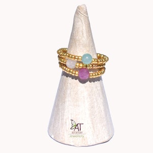 May include: A set of three gold beaded rings with a pink, white, and light blue gemstone. The rings are stacked on a white cone-shaped display stand.