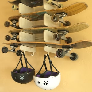 May include: A wall-mounted skateboard rack with five skateboards and two helmets hanging below. The skateboards are arranged in a stack, with the top board being the newest and the bottom board being the oldest. The helmets are black and white with a purple interior and a skull and crossbones logo.
