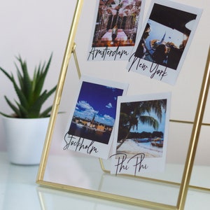 May include: A gold metal photo frame stand with four Polaroid photos inside. The photos are of various locations, including Amsterdam, New York, Stockholm, and Phi Phi.