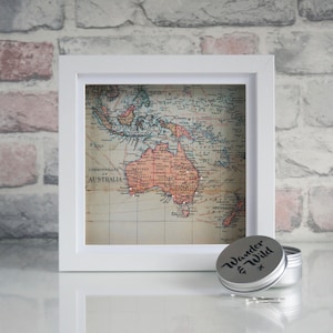May include: A white framed picture with a vintage map of Australia and surrounding islands. The map is decorated with small white pins. A silver tin with the text "Wander & Wild" is in the foreground.
