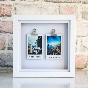May include: White picture frame with two photos of a city skyline. The photos are held in place with silver clips. The text "NEW YORK" and "19 OCT 2016" is printed below the photos.