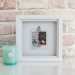 May include: A white framed print featuring a polaroid-style image of a Christmas tree in London, secured with a gold clip. The word "London" is written below. A light green candle is positioned to the left.
