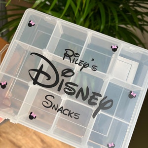 May include: A clear plastic storage box with multiple compartments. The box has a black and white Disney logo with the text "Do Riley's Disney Snacks". There are pink Minnie Mouse ears on each side of the box.
