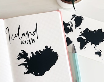 May include: A white notebook with a black outline of Iceland and the text "Iceland 02/09/19" on the left page. The right page shows a black outline of the world map.