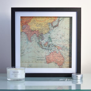 May include: A framed vintage map of Asia and Australia in a black frame with a white mount. The map is printed on paper with a light blue background and red, yellow, and brown markings. The map is displayed on a white surface.