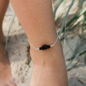 May include: A silver anklet with black beads on a woman's ankle. The anklet is on a sandy beach with green grass in the background. STERLING SILVER is written in the bottom left corner of the image.
