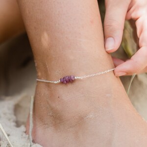 May include: A silver anklet with a row of small, purple gemstone beads. The anklet is on a person's ankle, with sand in the background.