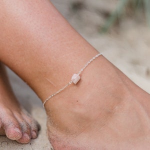 May include: A delicate silver anklet with a small, light pink gemstone charm. The anklet is shown on a person's ankle, resting on sand.