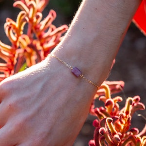May include: A delicate gold bracelet featuring a small, rectangular, purple gemstone. The bracelet is shown on a person's wrist against a backdrop of orange and red flowers.