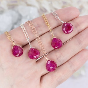 以下が含まれることがあります： 手の上に、ルビーのペンダントがついたネックレスが5本あります。ネックレスは、ゴールド、シルバー、ローズゴールドなど、さまざまなチェーンスタイルで作られています。
