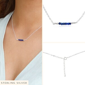 May include: A silver chain necklace with a row of small blue gemstone beads. The necklace is shown on a model wearing a light blue shirt. The necklace has an adjustable clasp.