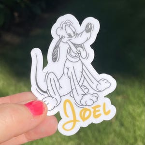 May include: A white sticker with a black and white line drawing of a cartoon dog. The dog is sitting with its tongue sticking out. The name "JOEL" is written in yellow cursive below the dog.
