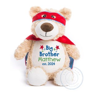 May include: A beige plush teddy bear wearing a red cape and a red and blue mask. The bear has a white patch on its chest with the embroidered text "Big Brother Matthew est. 2024" and red stars.