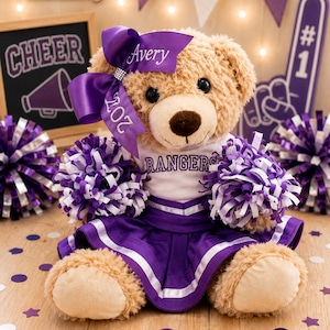 May include: A tan plush teddy bear wearing a white shirt with the word RANGERS in purple text and a purple pleated skirt with white trim. The bear has a large purple bow on its head with the name Avery and the year 2027, and holds two pom-poms.