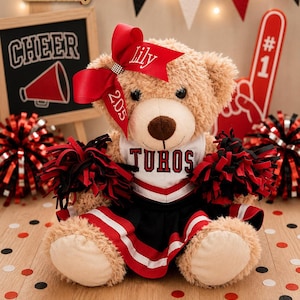 May include: A tan plush teddy bear dressed as a cheerleader, wearing a white and black uniform with the name TUROS embroidered on the chest. The bear has a large red hair bow with the name Lily and the number 205, and holds black and red pom-poms.