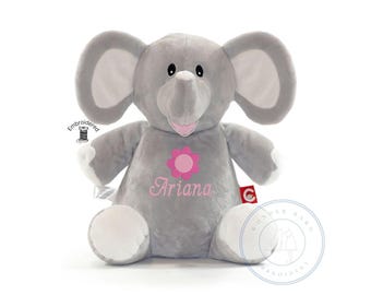May include: A soft, plush gray elephant toy with large ears, white accents on the feet, and a pink embroidered flower above the name Ariana in pink script. The toy features a small red tag on the side and a small embroidered spool icon.